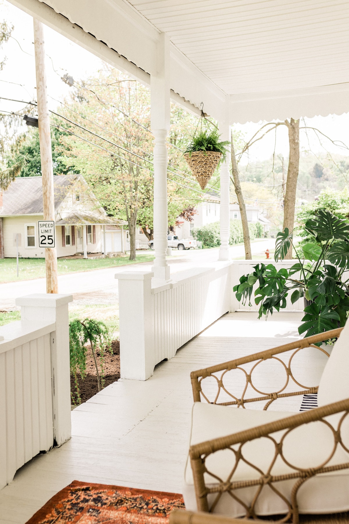 A welcoming front porch features a comfortable seating area with a rattan chair and a colorful rug. Lush greenery and a hanging planter are visible, enhancing the outdoor charm. A quiet street is seen in the background, lined with quaint houses and trees.