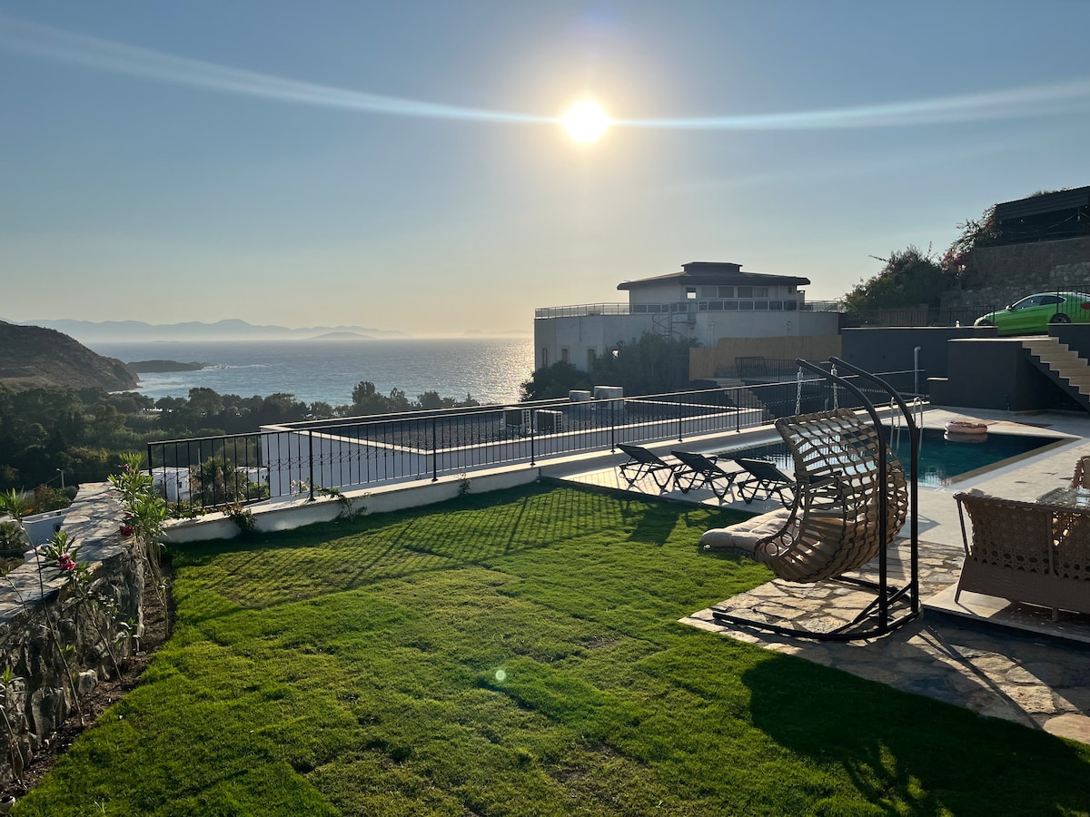 A scenic outdoor area features a private pool surrounded by lounge chairs on a manicured lawn. A comfortable seating arrangement provides a view of the sea, with sunlight reflecting off the water. The backdrop includes distant hills and a clear sky.