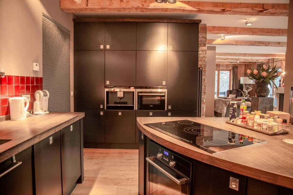 The kitchen area showcases a modern design with dark cabinetry and a curved wooden countertop. An induction cooktop is set into the island, complemented by built-in ovens. A variety of kitchen appliances and jars are arranged neatly, enhancing the functional layout.