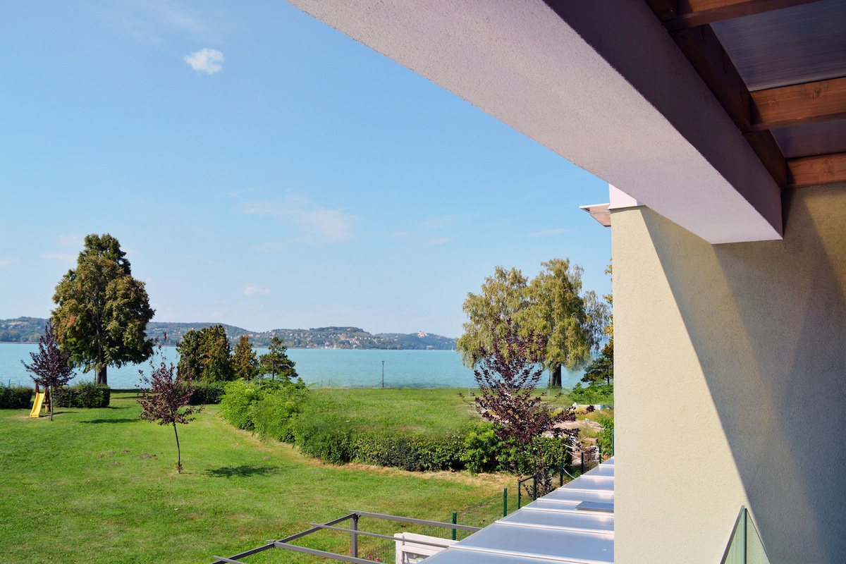 A scenic view of the lake is visible from the balcony, framed by lush greenery and trees. The peaceful shoreline is set against a clear blue sky, creating a tranquil outdoor space for relaxation.