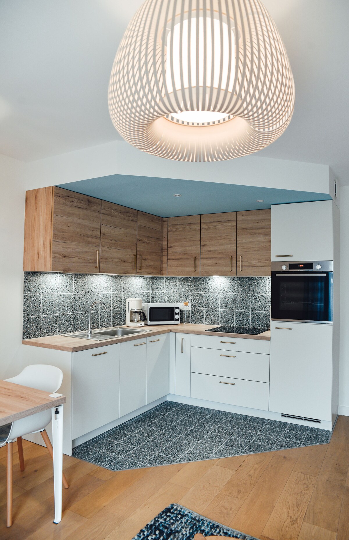 A modern kitchen is visible, featuring sleek white and wooden cabinetry. A dark geometric backsplash complements the light countertops. Appliances include a microwave and an oven. A wooden dining table with white chairs is positioned nearby, illuminated by a woven pendant light above.