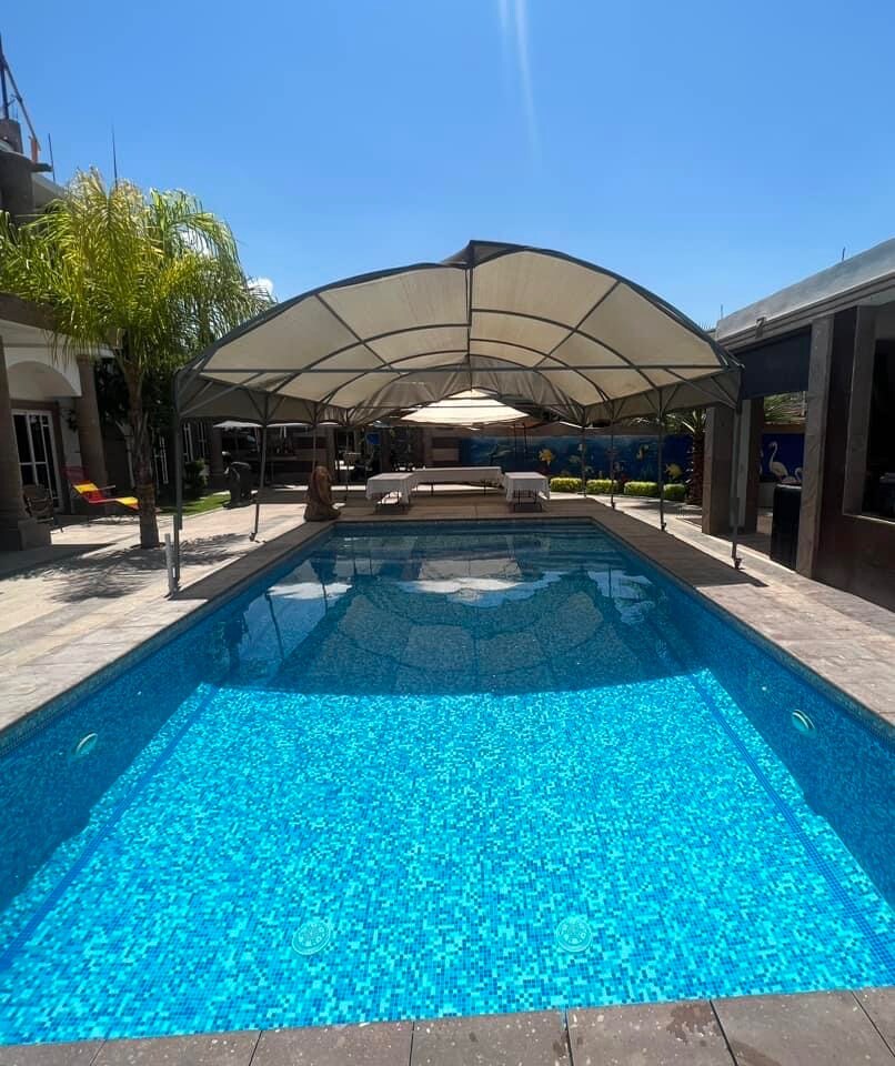 A spacious outdoor pool area is bordered by lush greenery, featuring a covered seating area with comfortable lounge furniture. The inviting, clear blue water reflects sunlight, enhancing the tranquil setting, perfect for relaxation and family enjoyment.