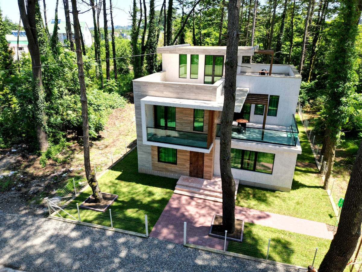A modern three-story structure is surrounded by lush greenery, showcasing large windows and glass balconies. A pathway leads to the main entrance, bordered by a grassy area and trees, contributing to the serene setting of the property.