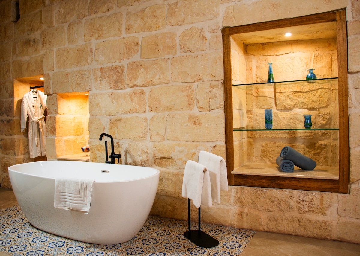 A soothing bathroom space features a freestanding white bathtub beside a tiled floor adorned with intricate patterns. Natural stone walls create a warm ambiance. A wooden shelf displays decorative items, while neatly rolled towels are placed beside the tub, adding to the room's functionality.