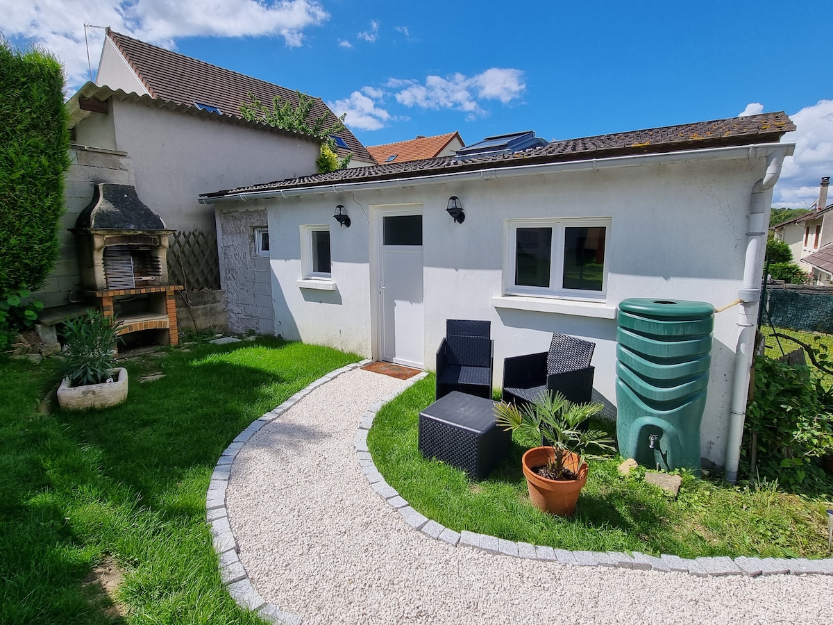 A garden area features a stone pathway leading to a compact dwelling. Black patio chairs are arranged near a potted plant, accompanied by a water tank and a barbecue grill positioned to the side. Lush greenery adds to the inviting outdoor space.