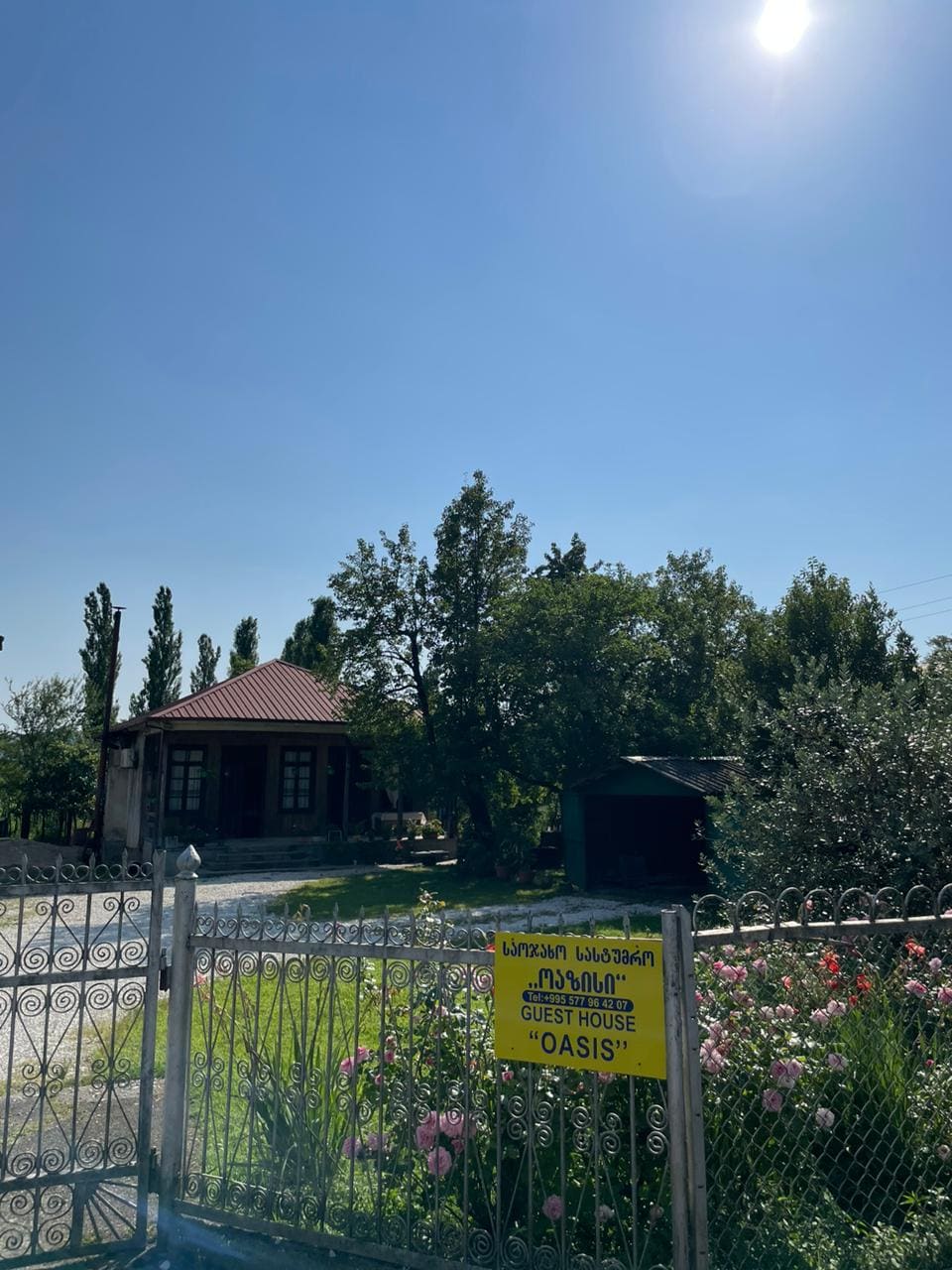 A welcoming guest house named 'OASIS' is displayed on a bright yellow sign at the entrance. The house, featuring a red roof, is surrounded by lush greenery and blooming flowers, under a clear blue sky. A well-maintained garden adds to the inviting atmosphere.