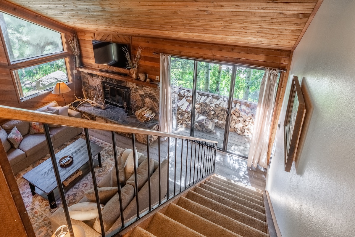 A welcoming living area is visible with warm wood paneling and a stone fireplace. Large glass doors lead to an outdoor space, allowing natural light to fill the room. A comfortable sofa and coffee table are arranged for relaxation, with decor elements enhancing the cozy ambiance.