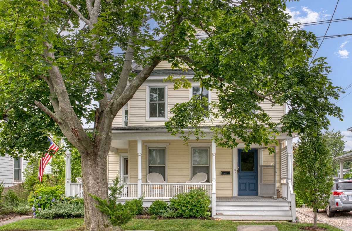The exterior of a charming historic home is displayed, featuring a welcoming front porch with two chairs. A large tree provides shade, while an American flag is seen hanging. The house is painted in light colors, complementing the green of the surrounding landscaping.