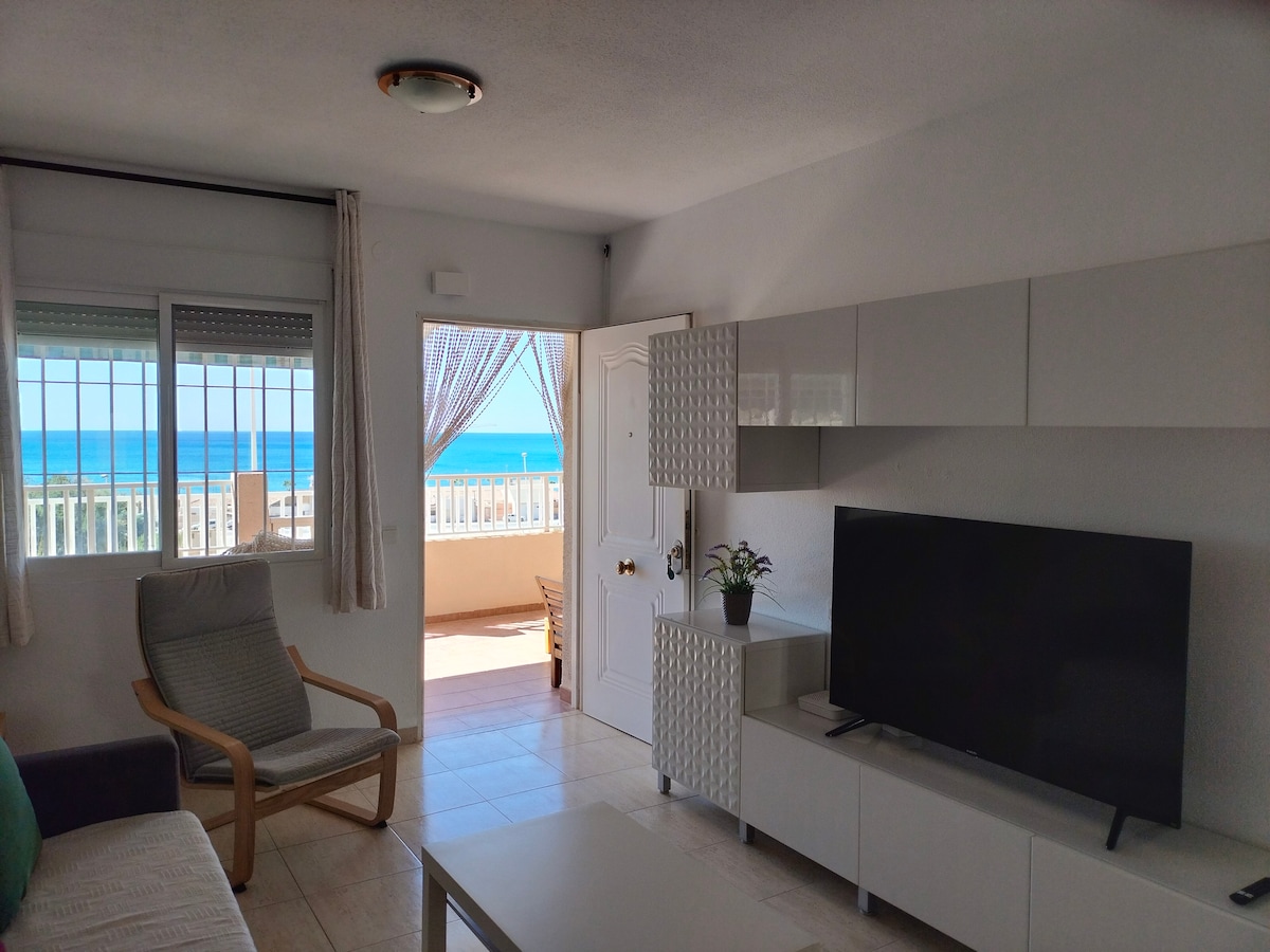 A bright living area features a comfortable chair and a modern television unit. Natural light floods in from the open door, framing a scenic view of the sea through a balcony. Sheer curtains gently sway in the breeze, enhancing the inviting atmosphere.