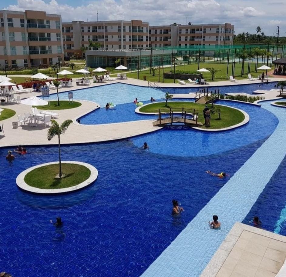 The image showcases a large pool area with a winding design. Guests can be seen enjoying the water, surrounded by palm trees and lounge chairs. Multiple green spaces and a bridge are visible, along with the condominium buildings in the background.