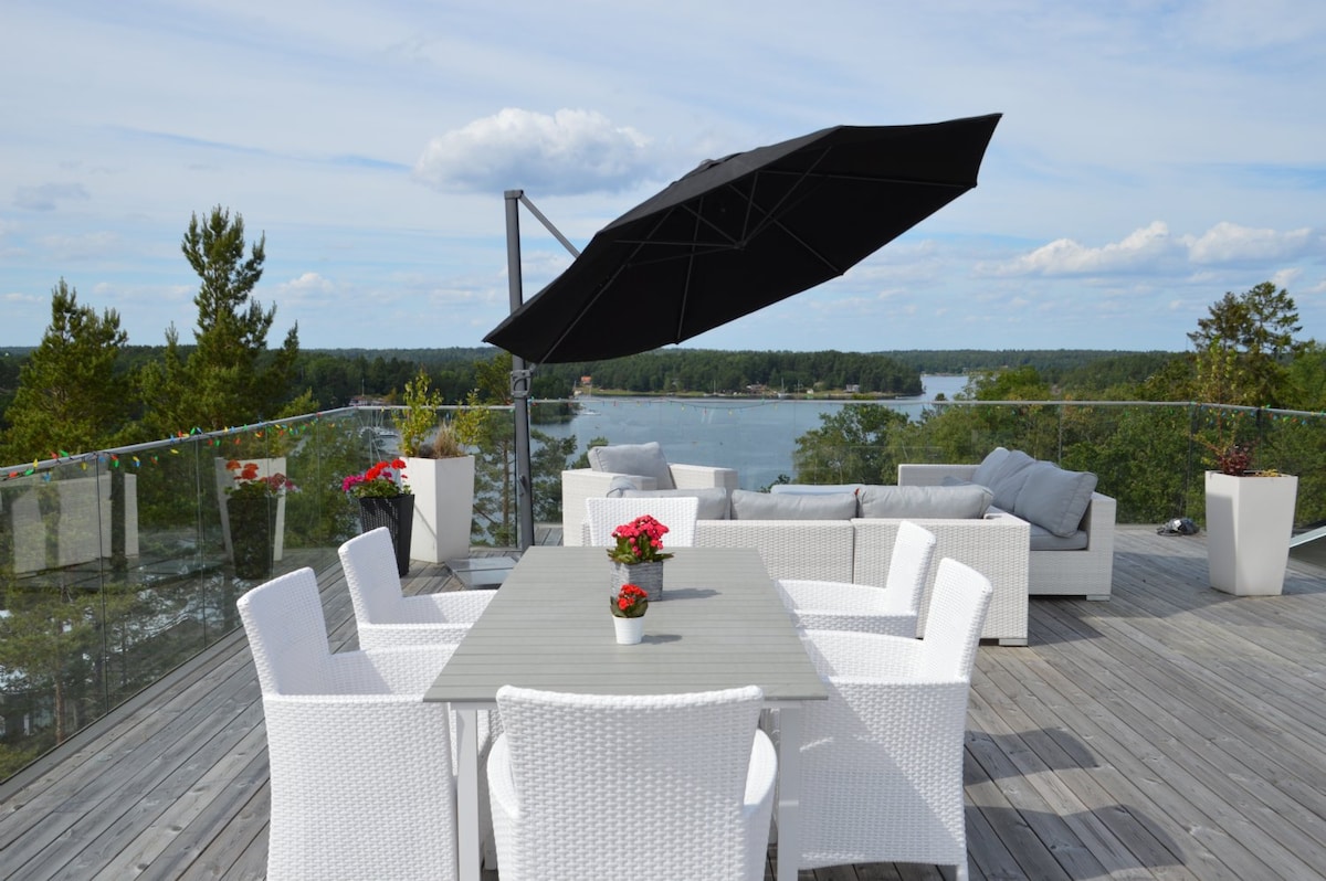 An outdoor terrace features a spacious dining table surrounded by white wicker chairs. A large black umbrella provides shade, while potted flowers add a touch of color. The area offers scenic views of the water and surrounding greenery.