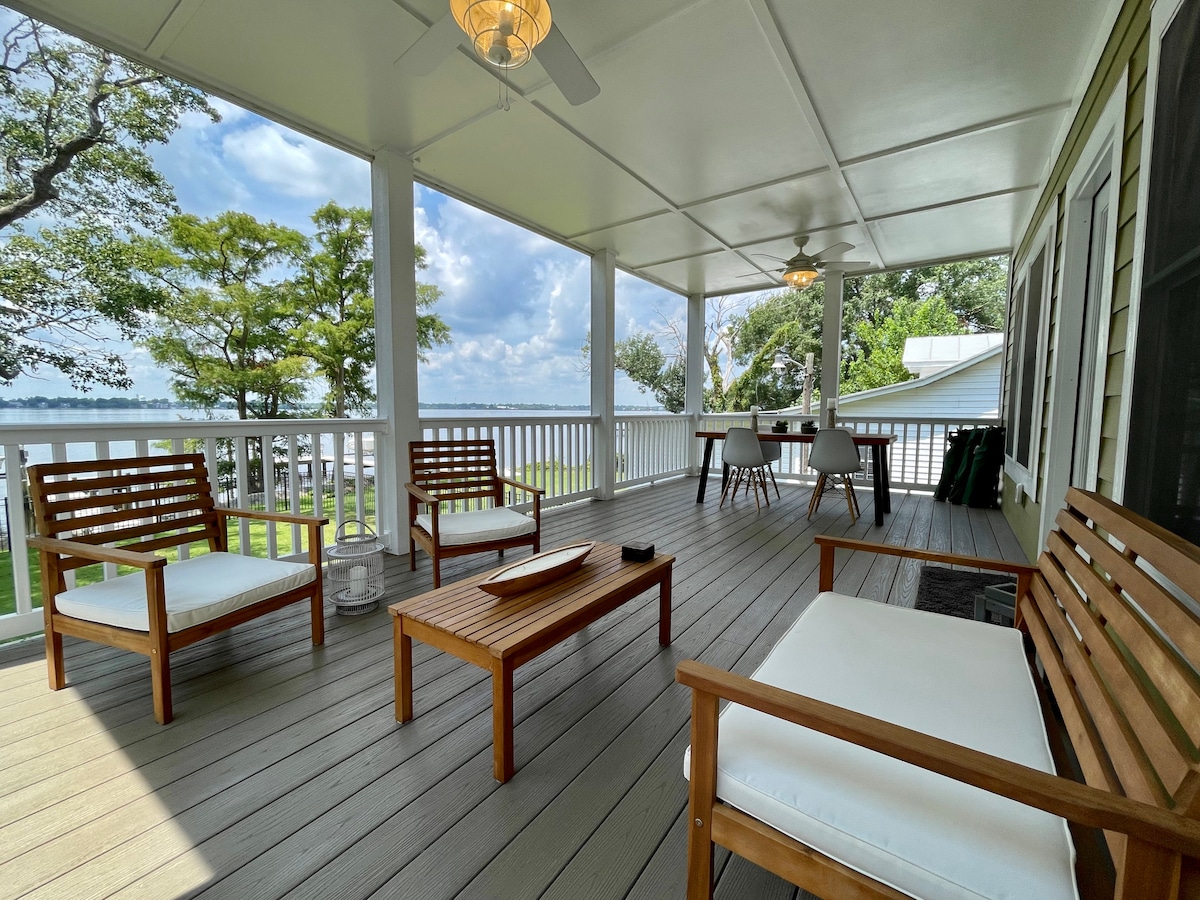 A spacious screened porch features wooden seating arrangements, providing a comfortable area for relaxation. Light-colored cushions are set on the chairs and sofas. A dining table is positioned in the background, with water views visible through the open railings.