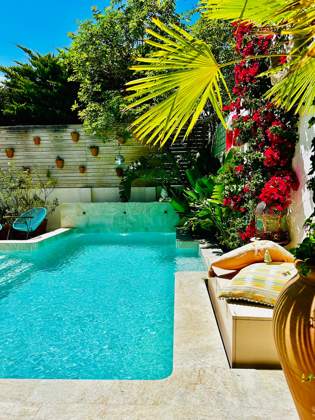 A serene patio garden is featured, complete with a turquoise pool surrounded by lush greenery and vibrant flowers. A comfortable lounge area with decorative cushions is positioned nearby, inviting relaxation under the warm sunlight.