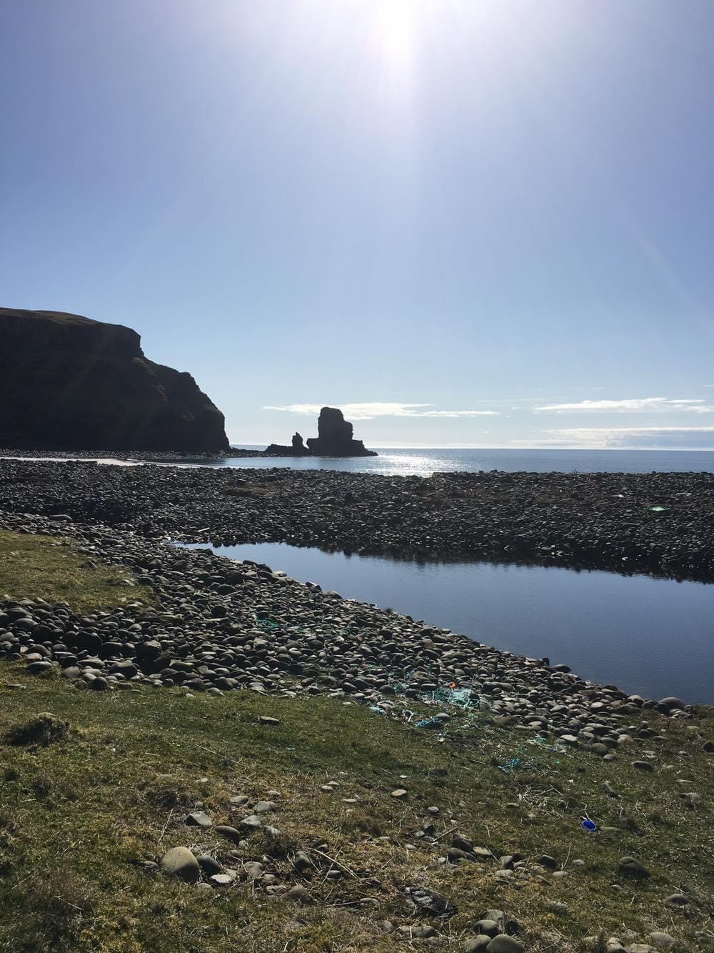 A rocky shoreline is bathed in sunlight, featuring smooth pebbles scattered along the water's edge. The tranquil sea reflects the bright sky, with rugged cliffs rising in the background. A distant rock formation is visible offshore, creating an interesting focal point in the natural landscape.