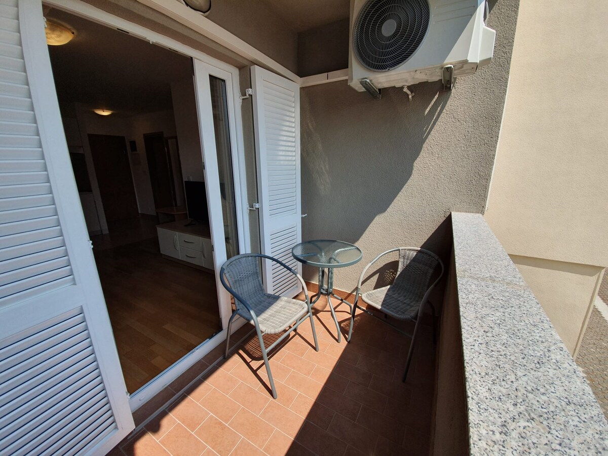 A small balcony is featured, with two rattan chairs and a circular glass table positioned on tiled flooring. White shutters are opened, allowing access to the interior space that is softly lit, while an air conditioning unit is visible on the wall above.
