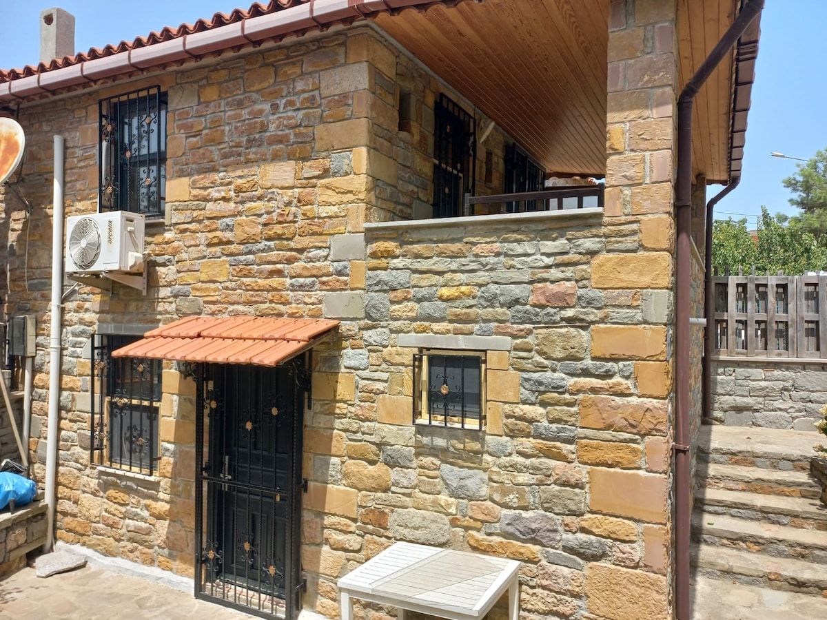 A stone house with a sloped roof is featured, showcasing a blend of textured stones in earthy tones. Black window grilles provide security, and an air conditioning unit is mounted on the exterior. A small outdoor table is positioned in front of the house.
