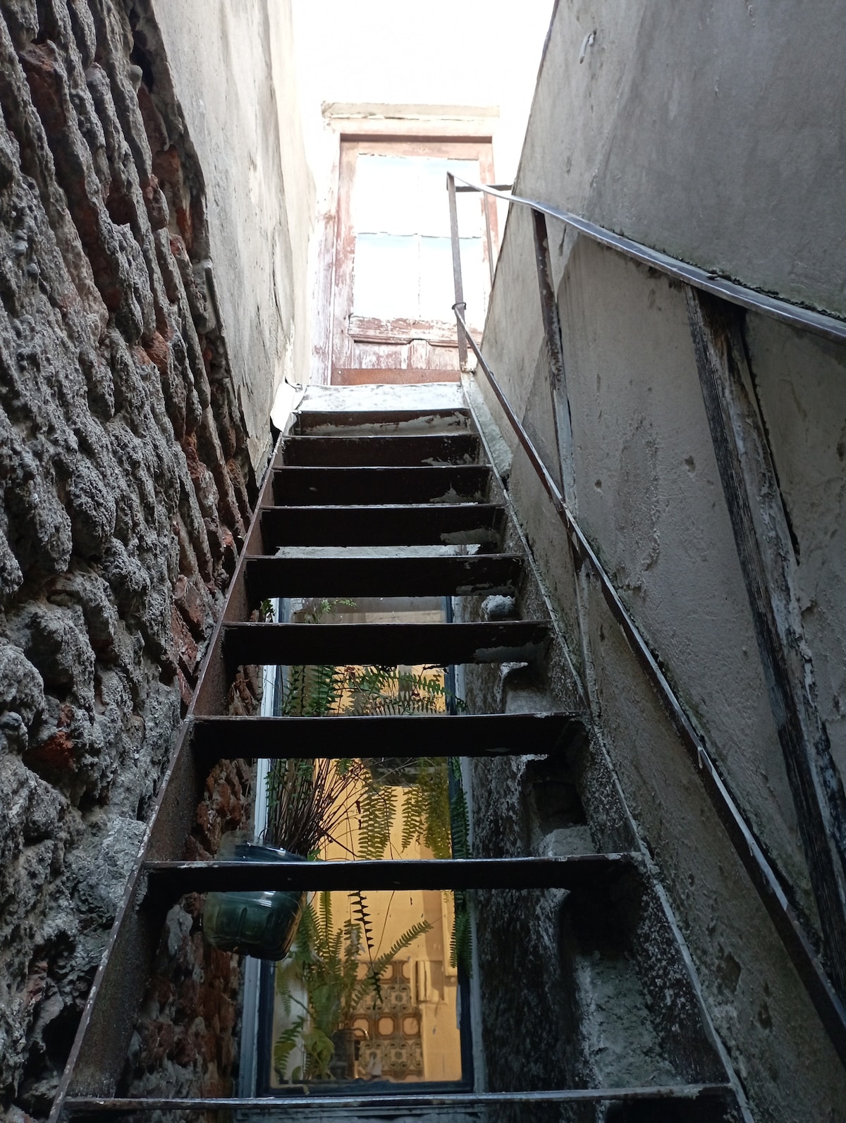 A narrow staircase with metal and stone steps ascends toward a weathered wooden door, allowing natural light to illuminate the space. An indoor plant is visible through a window beside the staircase, contributing to the serene atmosphere.