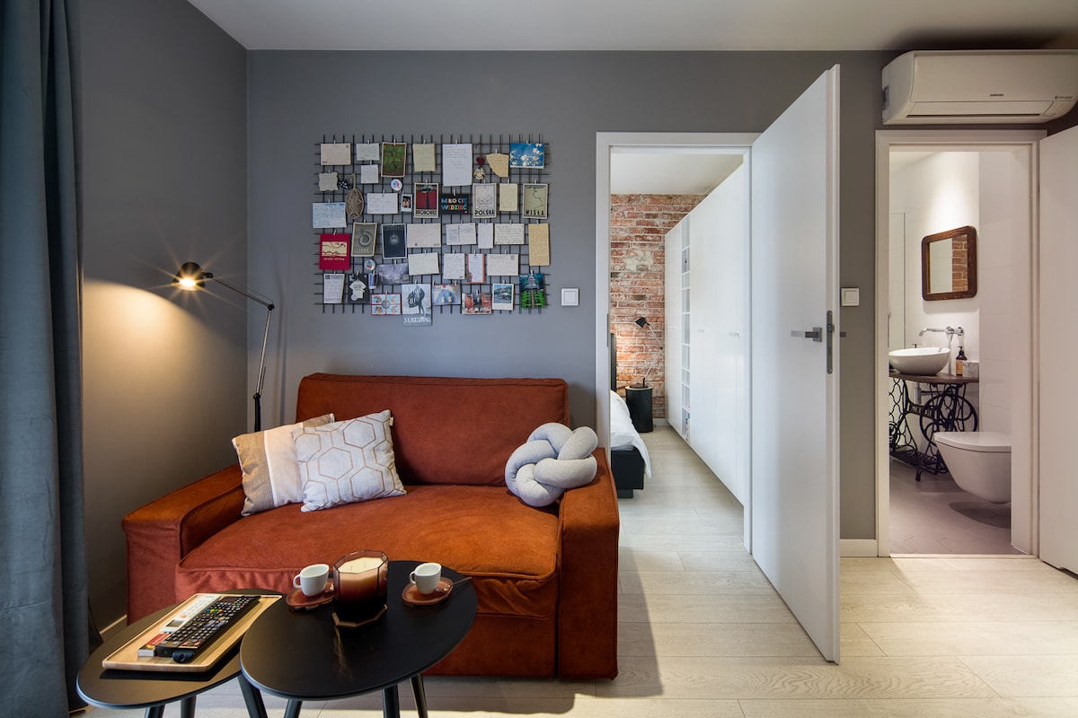 A cozy seating area features a brown sofa adorned with pillows, positioned next to a small black table with two cups. A gallery wall of framed art pieces adds visual interest. The doorway leads to a glimpse of the hallway and bathroom beyond.