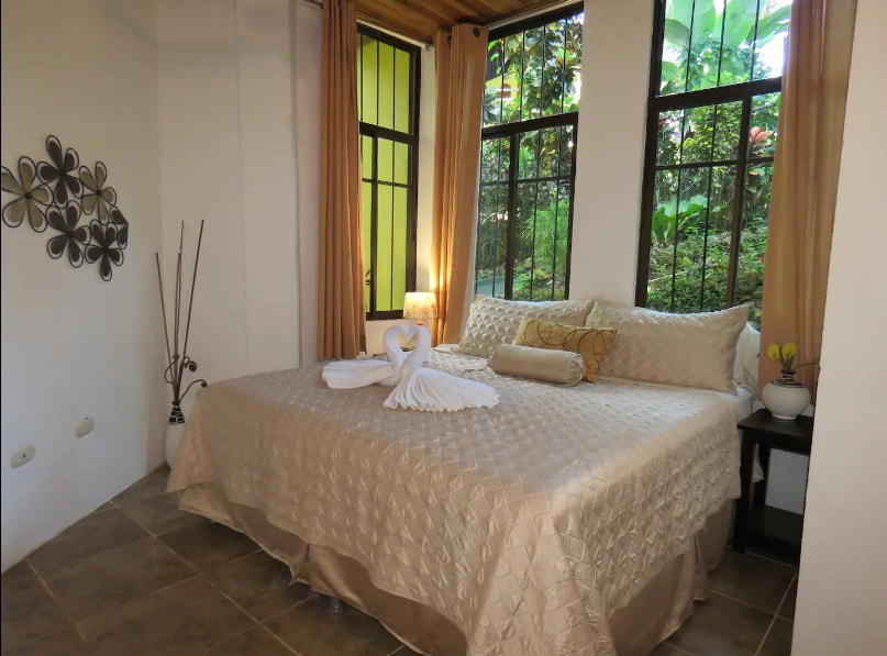 A spacious bedroom is furnished with a large bed, adorned with neutral bedding and decorative pillows. Natural light fills the room through tall windows, which overlook lush greenery. A bedside table holds a small lamp, and decorative elements enhance the calming atmosphere.