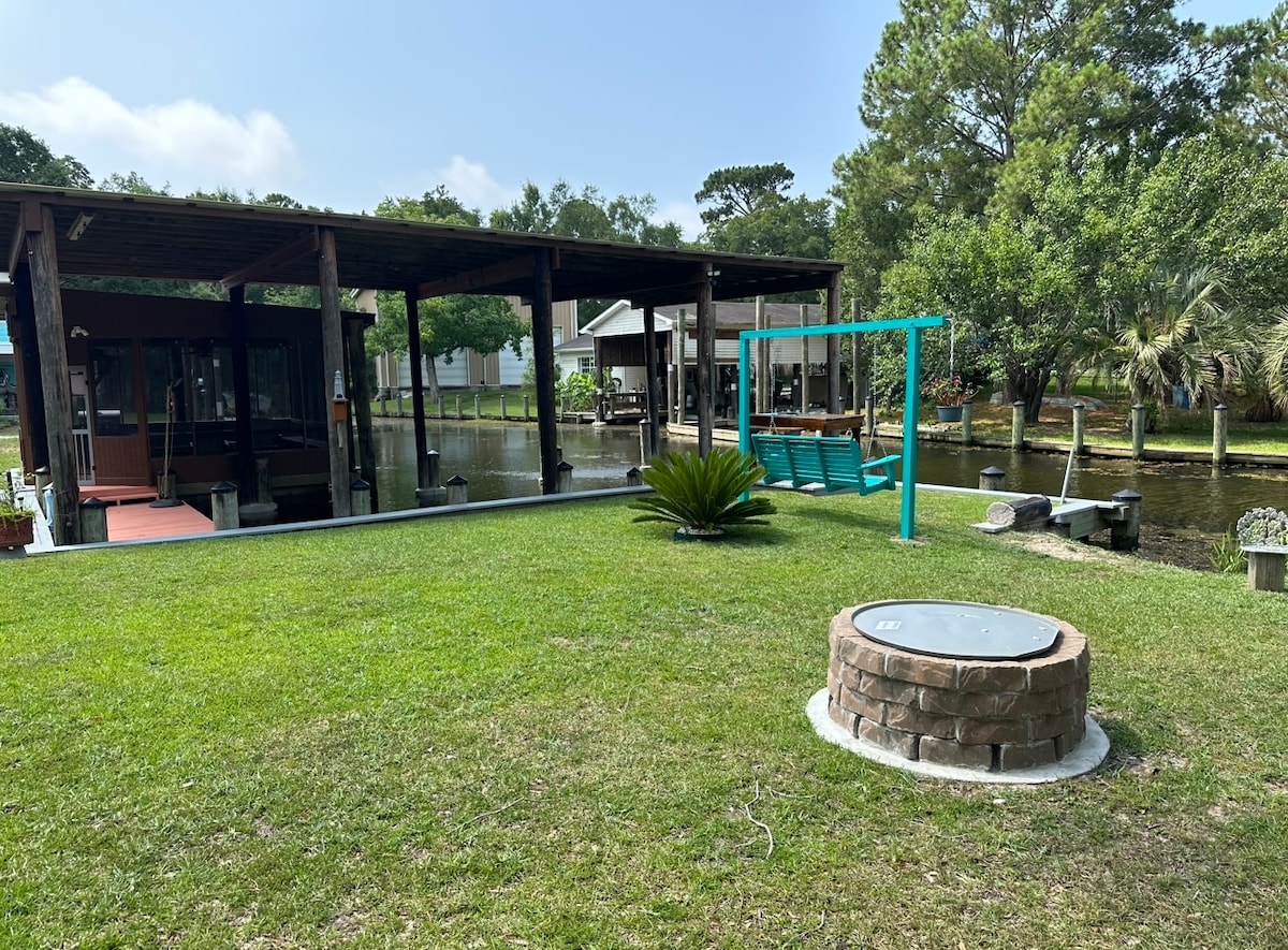 An outdoor area is depicted featuring a manicured lawn leading to a covered boathouse. A swing hangs nearby, and a stone fire pit is visible on the ground. Surrounding greenery and water provide a serene backdrop.
