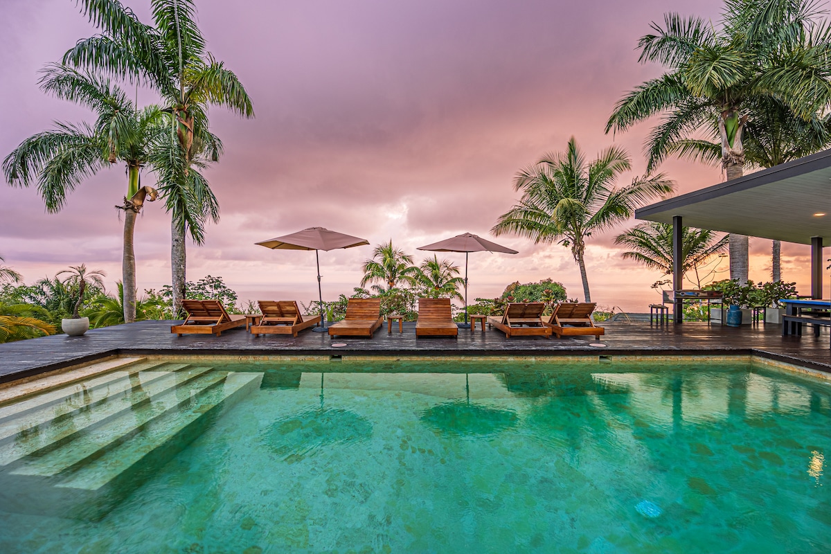 A tranquil pool area is surrounded by palm trees and lounge chairs overlooking the ocean. Umbrellas provide shade, and wooden decking frames the space. The sky is filled with a blend of soft pink and gray hues, reflecting the serene atmosphere of the setting.