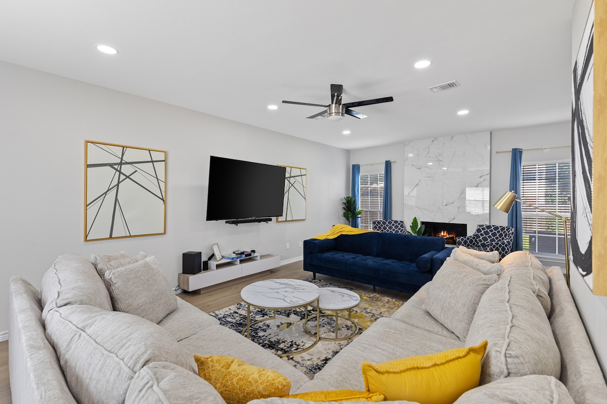 A spacious living area features a large sectional sofa in light gray and a blue couch, arranged around a round coffee table. Wall-mounted smart TV is positioned above a media console. Bright accents are provided by artwork and curtains, complementing the neutral decor.