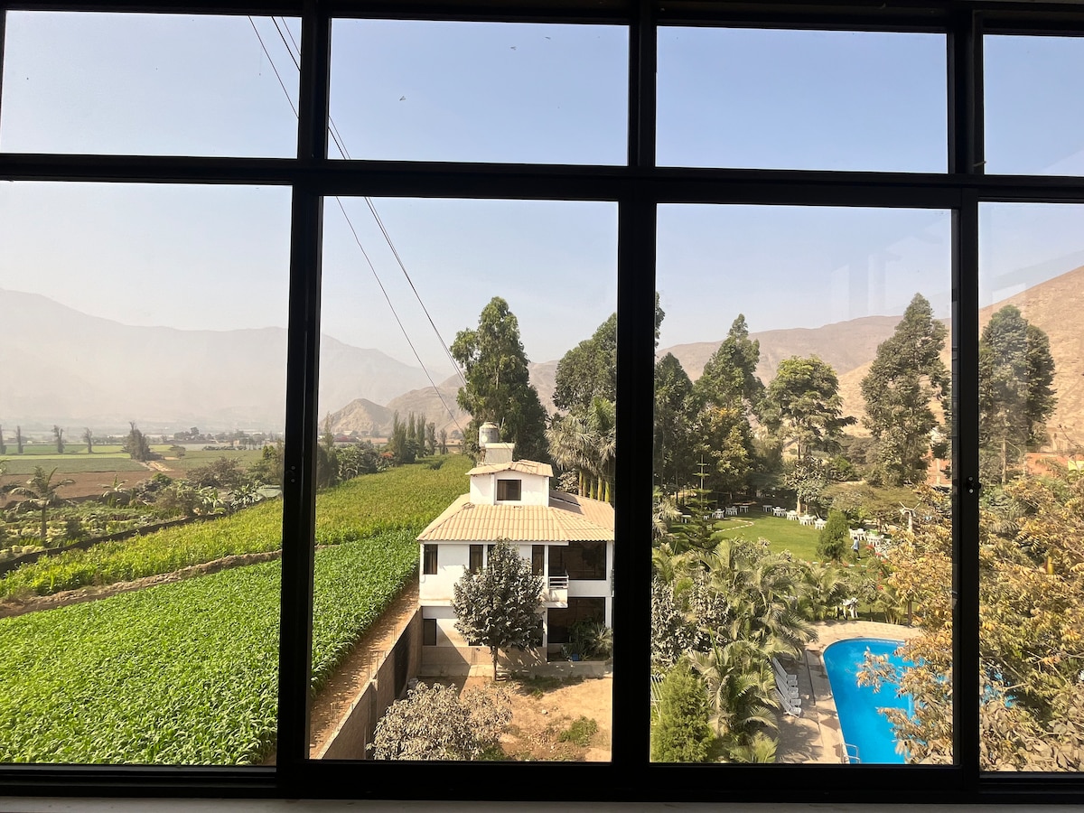 A panoramic view is captured through large windows, showcasing lush fields and rolling hills in the distance. The image includes a glimpse of the pool area and the nearby family-friendly lodge surrounded by greenery.