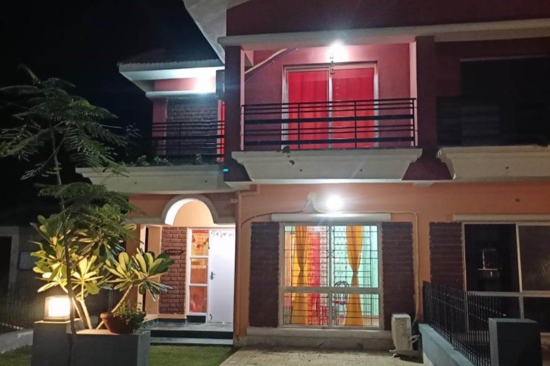 The two-story bungalow is shown at night, highlighting illuminated entry points and vibrant colored curtains visible through the windows. A lush green plant is positioned near the entrance, while outdoor lighting enhances the inviting ambiance of the façade.
