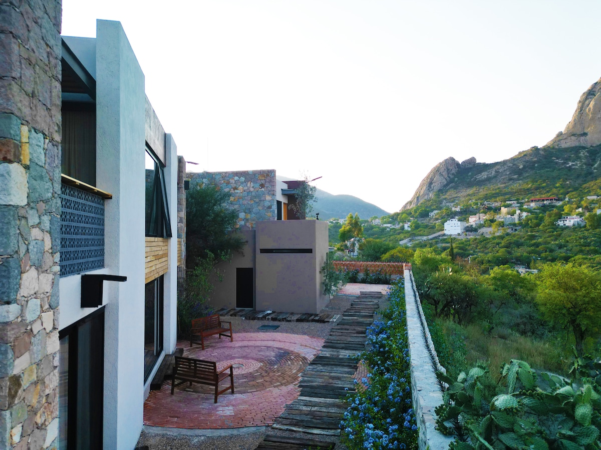 A minimalistic outdoor space is surrounded by stone and wood structures, featuring a circular seating area made of natural materials. Lush greenery and blooming flowers add color to the landscape. Views of the surrounding mountains provide a scenic backdrop.