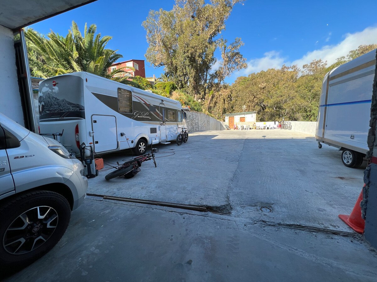 A spacious parking area is offered for camper vans and recreational vehicles, surrounded by greenery and tall trees. A mix of parked vehicles, including a camper van and a bicycle, can be seen on the smooth concrete surface under a clear blue sky.