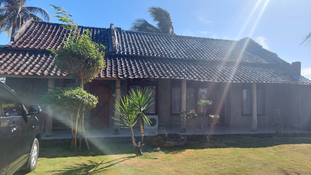 A welcoming exterior view of the property is presented, featuring a traditional roof with a blend of stone and wood accents. Lush greenery surrounds the entrance, and a vehicle is parked nearby. Sunlight streams through the foliage, creating a serene atmosphere.