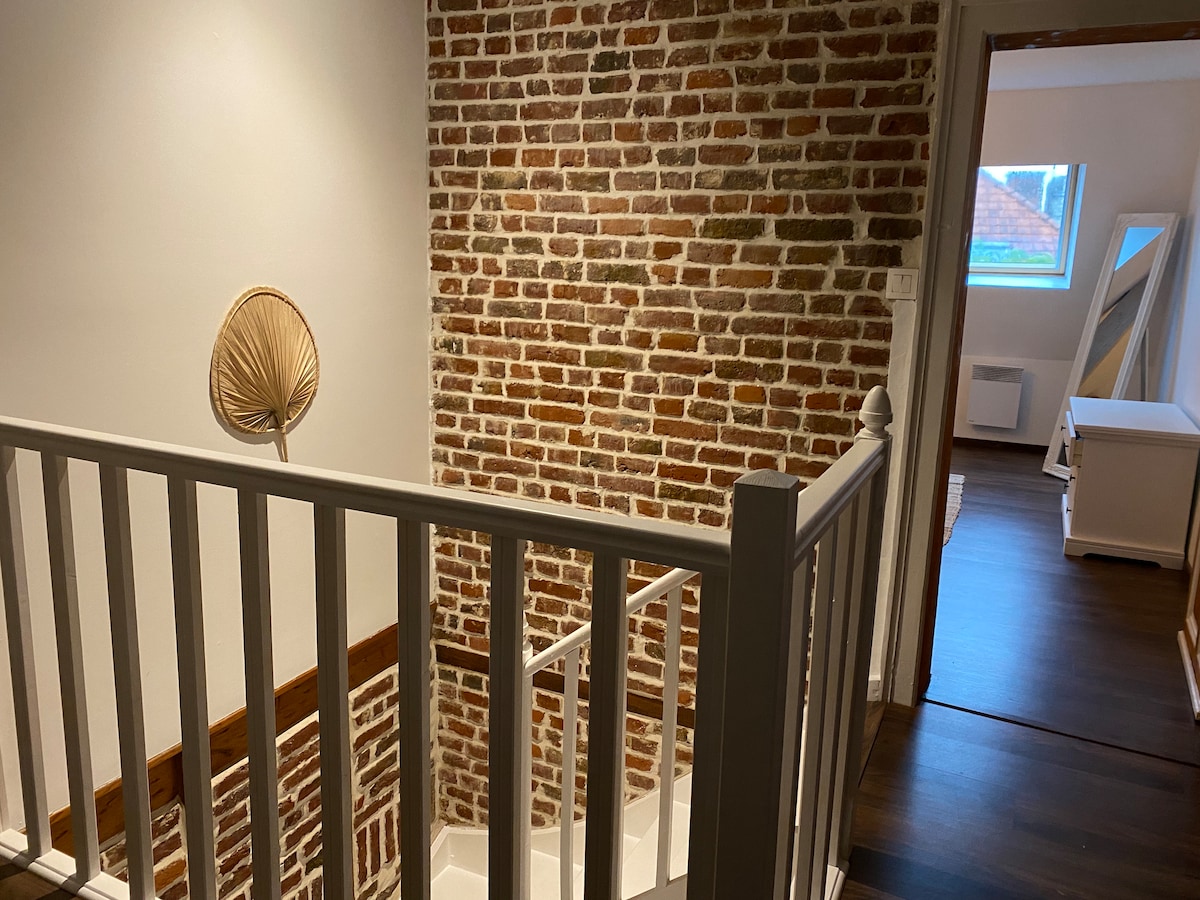 A staircase with white railings provides access to the upper level, framed by a textured brick wall. Natural light filters through a window, illuminating the warm wooden floor and leading to a well-lit room beyond.