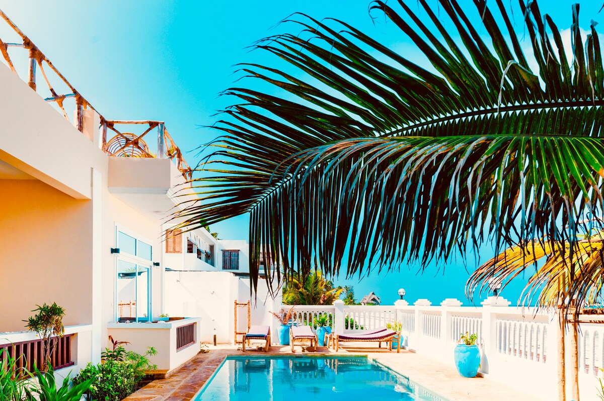 A serene outdoor area features a refreshing pool surrounded by tropical plants and lounge chairs. A tall palm tree provides shade, while the clear blue sky adds to the tranquil atmosphere. The white walls and accents echo a coastal vibe.