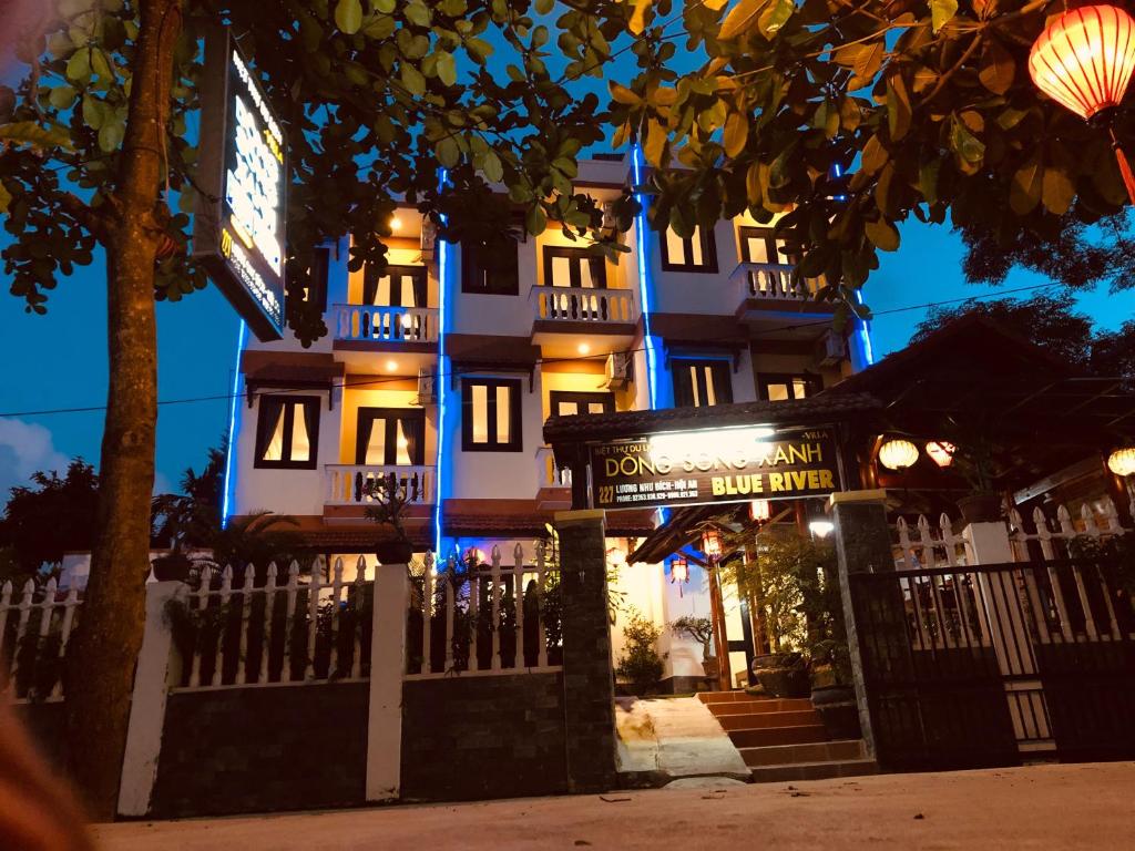 The exterior of the villa is showcased with illuminated signage and multiple floors adorned with blue lighting. A welcoming entrance is accentuated by plants and lanterns, while a pathway leads towards the door. The surrounding area includes white fencing and well-maintained greenery.