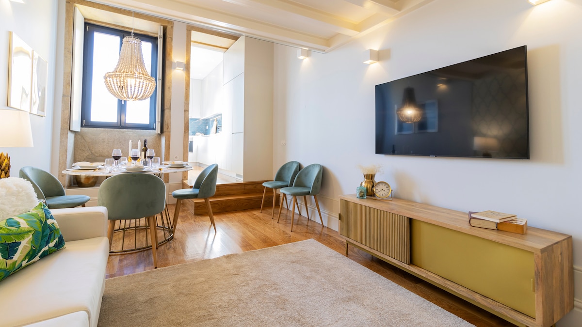 A light-filled living area features a comfortable sofa and a large television mounted on the wall. A dining area with modern chairs is visible in the background. Warm wooden floors complement the elegant lighting fixtures, enhancing the room's spacious feel.