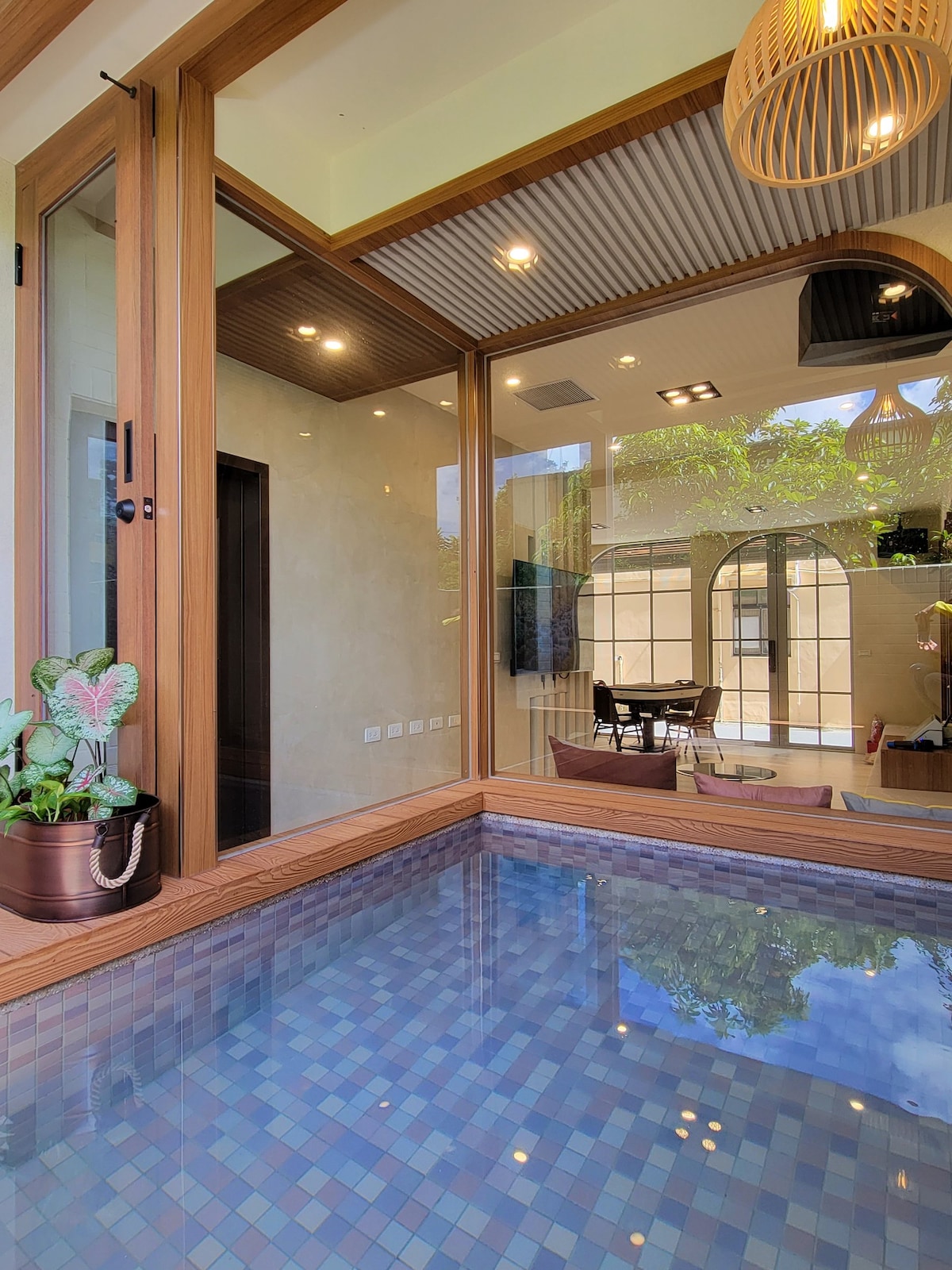 A serene indoor swimming pool is surrounded by wooden framing and large glass panels. The tiled water features a mosaic pattern. Soft lighting illuminates the space, while a potted plant adds a touch of greenery. A dining area is visible through the glass, enhancing the open layout.