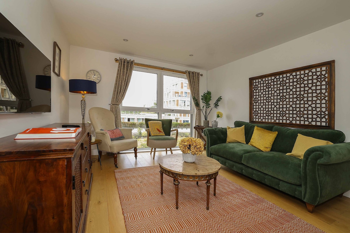 The open plan living room is highlighted by a large green sofa with yellow cushions. A wooden coffee table sits centrally, topped with a floral arrangement. Natural light fills the space through large windows, with a view of the communal garden visible outside.