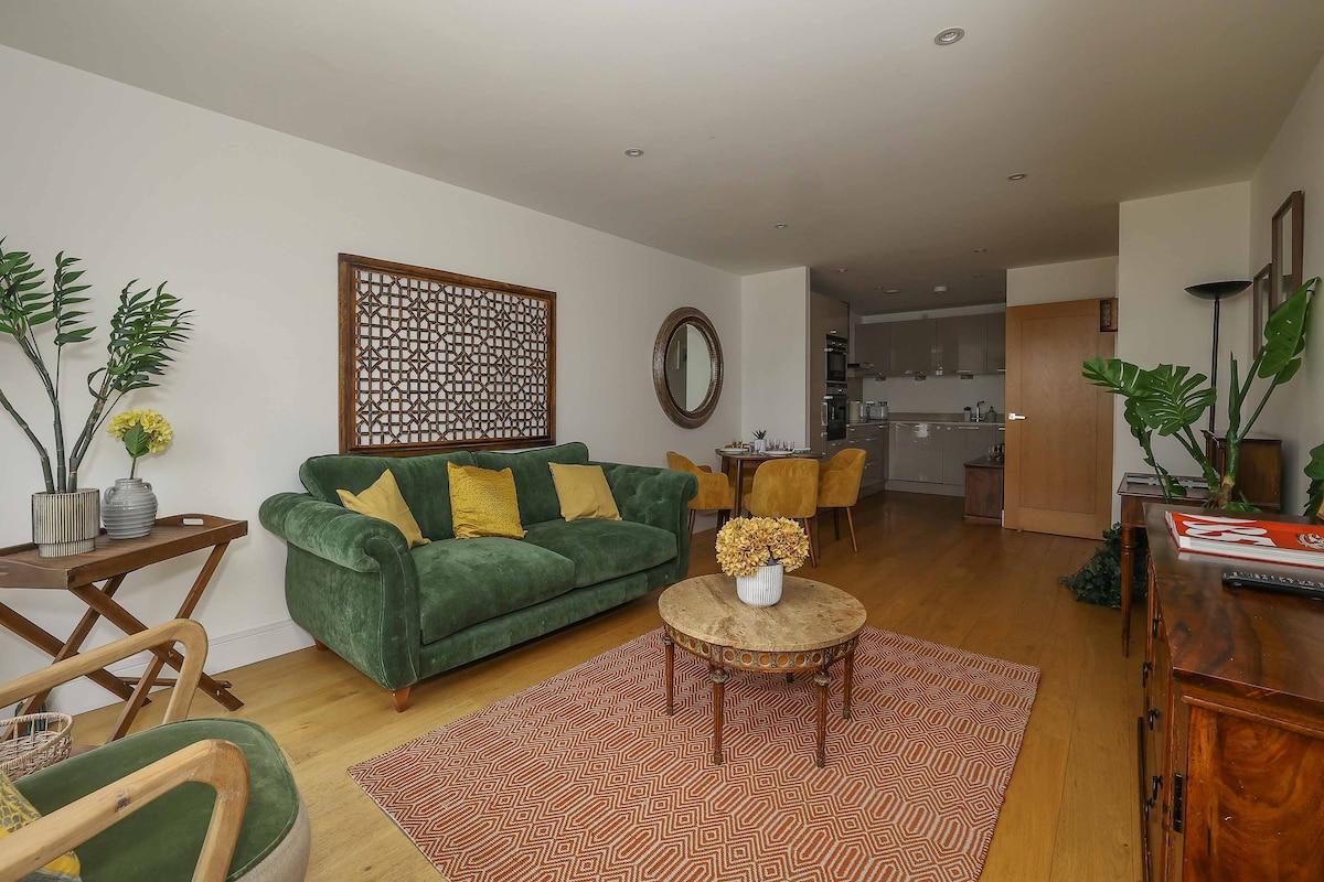 A spacious living area showcases a green, comfortable sofa accented by yellow cushions. A round coffee table stands on a textured rug, surrounded by tasteful decor elements. In the background, an open-plan kitchen area is visible, enhancing the inviting and practical design of the space.