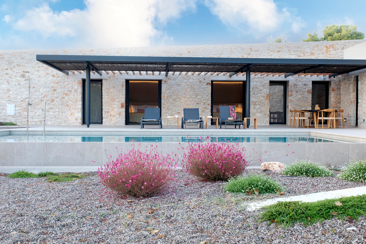 A modern villa is shown with natural stone walls, framed by a spacious terrace. Sun loungers are positioned near the pool, surrounded by lush garden beds with blooming flowers. Sliding doors provide access to the interior dining area, enhancing the indoor-outdoor connection.