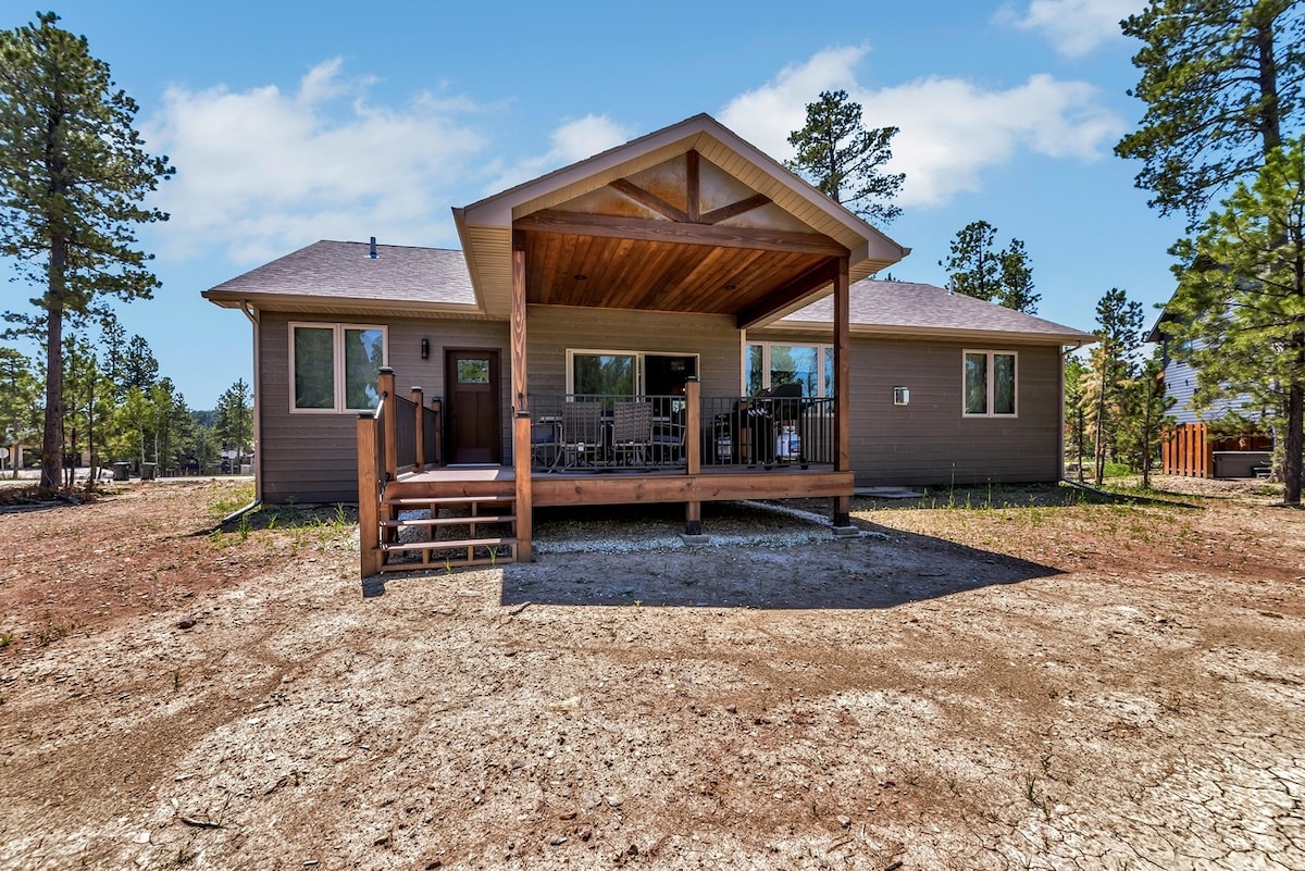 A spacious house is set against a backdrop of tall trees, featuring a welcoming porch with wooden accents. The outdoor area showcases ample space for relaxation, with a pathway leading to the entrance. Large windows reflect natural light, enhancing the home's inviting aspect.