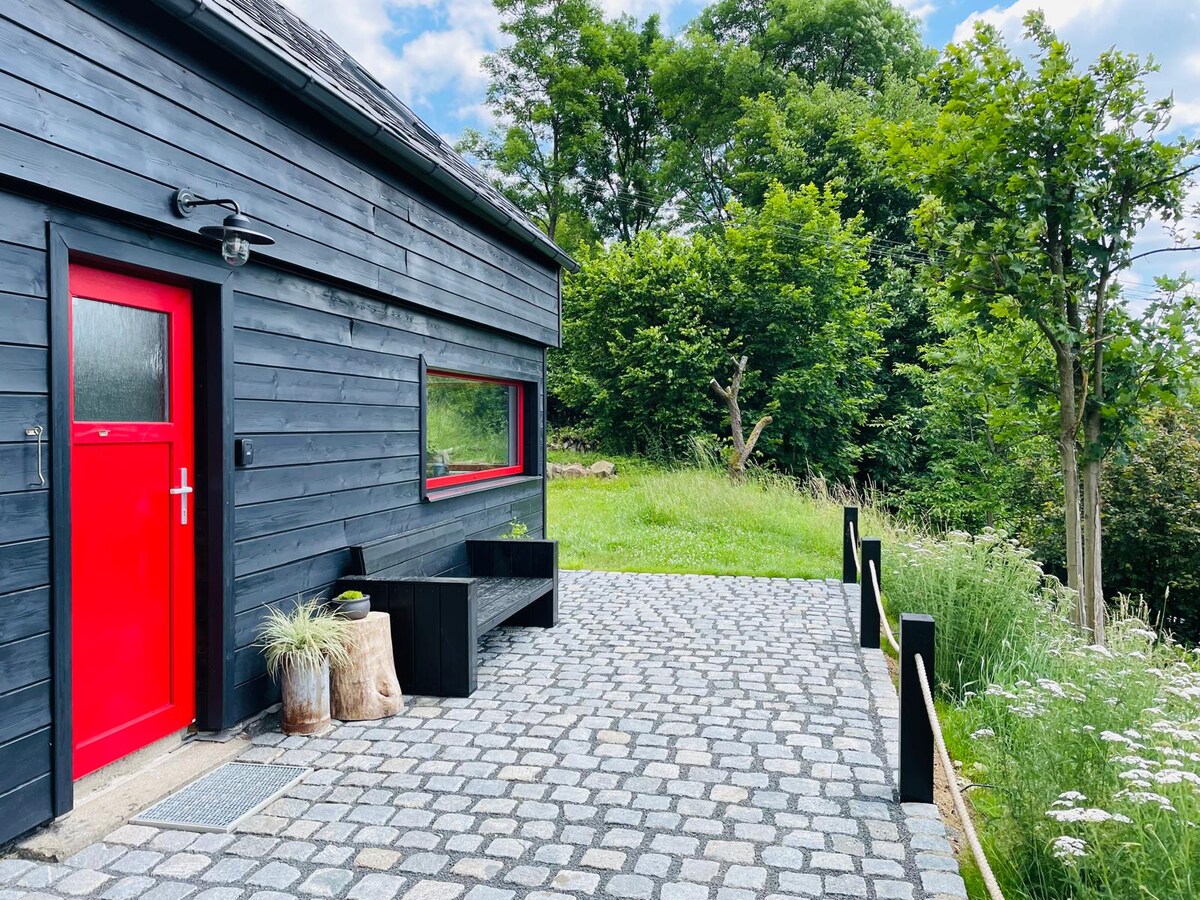 A black wooden exterior is complemented by a vibrant red door and a large window, providing a striking contrast. The cobblestone patio features a simple bench and natural elements, while lush greenery surrounds the area, creating a serene outdoor space.