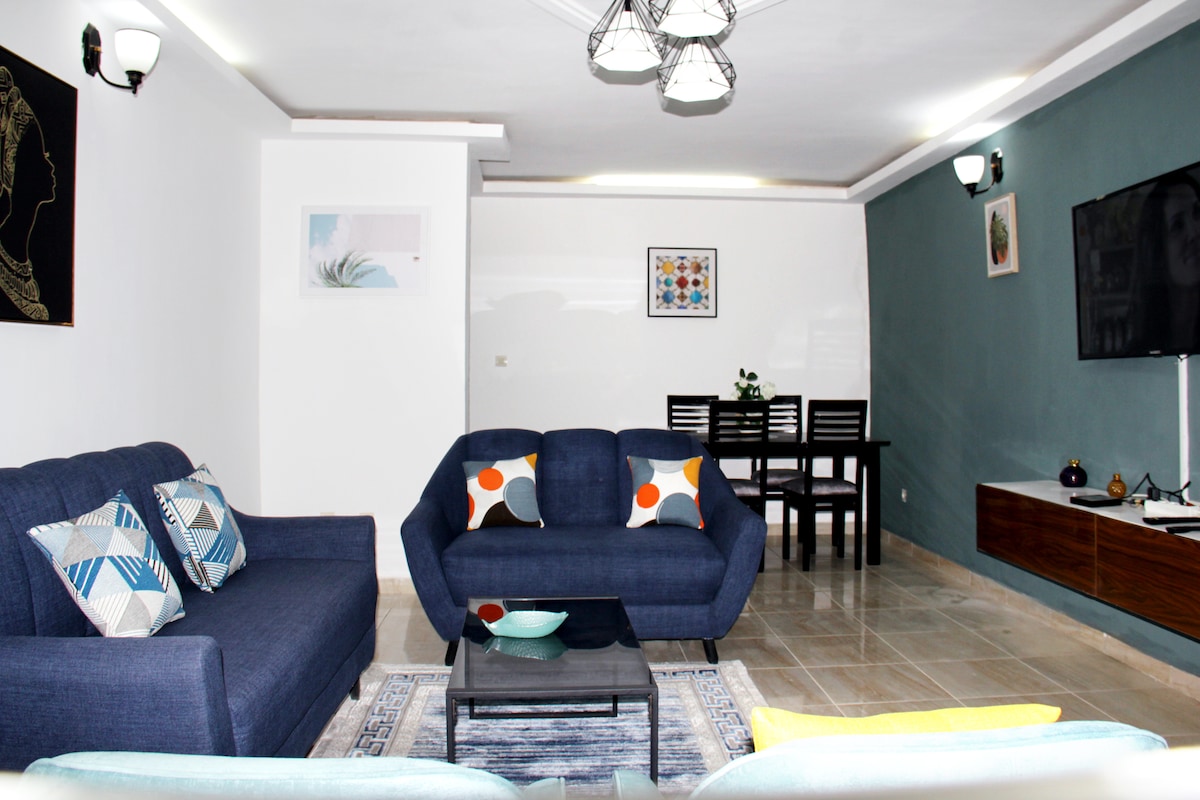 A spacious living area is equipped with a comfortable blue sofa and a complementary armchair adorned with colorful cushions. A rectangular coffee table sits at the center, and a dining table for six is positioned against the wall. A flat-screen TV is mounted on a media console.
