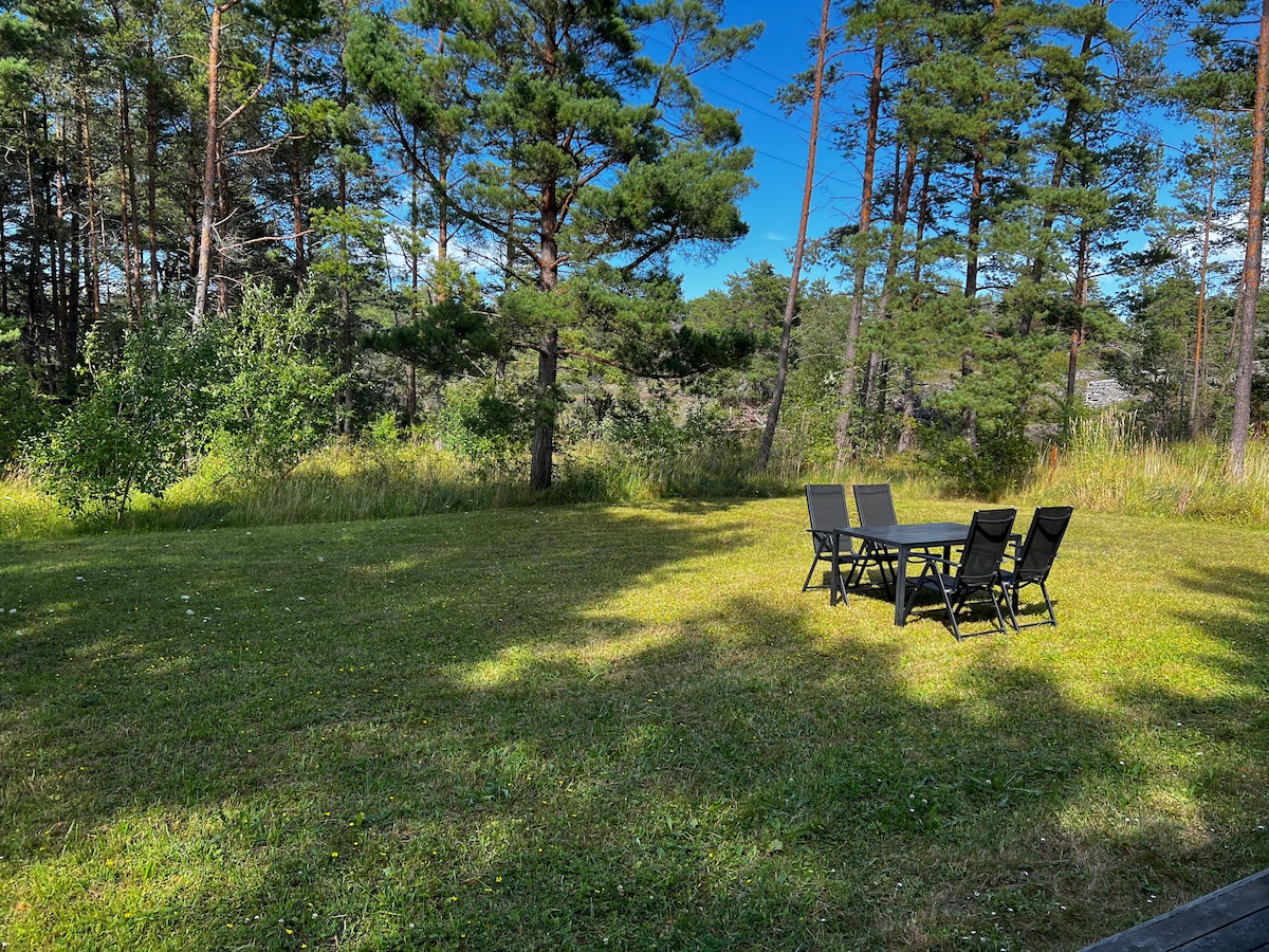 A spacious outdoor area is surrounded by tall trees and lush greenery. A black dining set with six chairs is positioned on a manicured lawn, creating an inviting space for meals and gatherings. Sunshine filters through the leaves, casting gentle shadows on the ground.