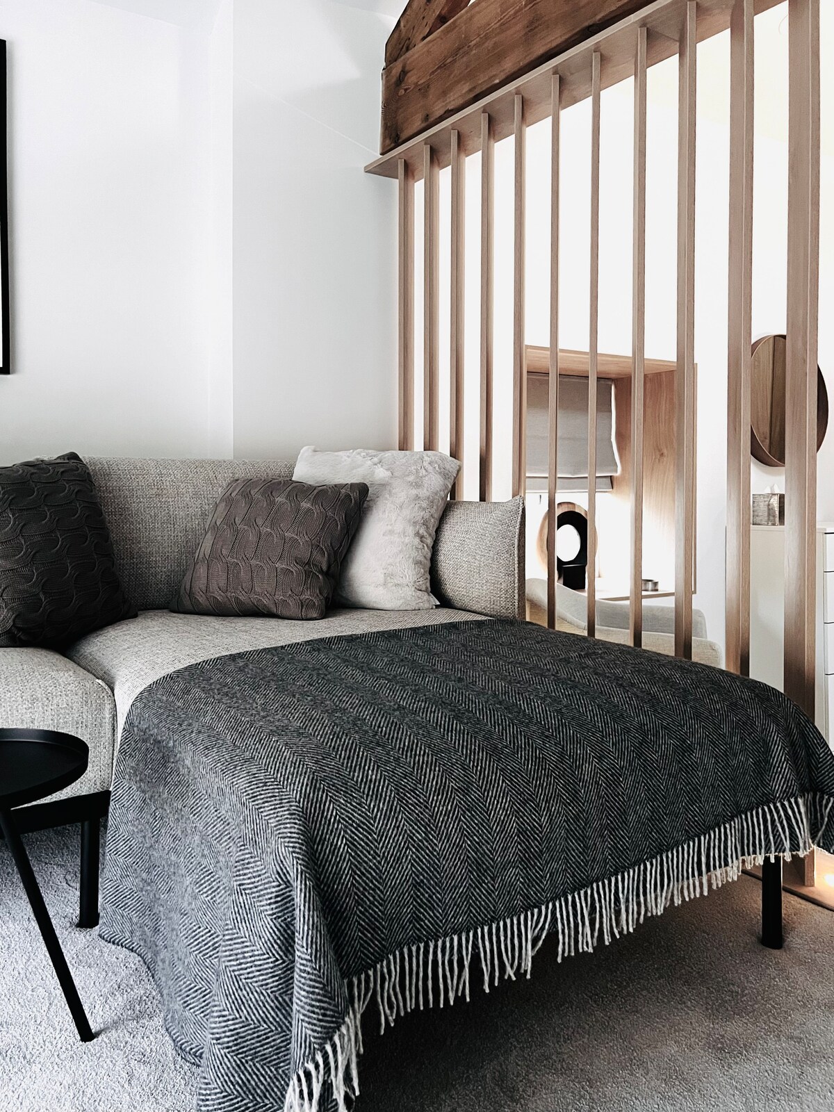 A comfortable seating area is featured, showcasing a spacious sofa adorned with textured cushions. A dark, round table is positioned nearby. A soft, fringed throw is layered over the seat, contributing to the inviting design. Natural light enhances the neutral tones within the space.
