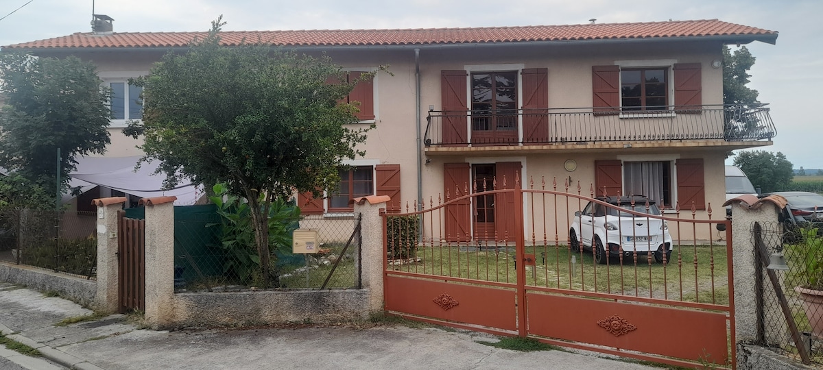 The exterior of a two-story house is featured with a light-colored facade and brown shutters. A gated entrance leads to a grassy yard, where a white vehicle is parked. Tree foliage and outdoor seating areas can be seen, creating an inviting atmosphere.