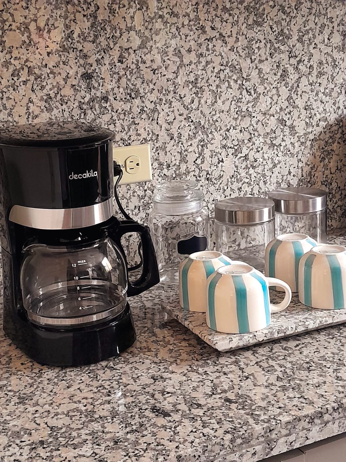 A kitchen countertop features a modern coffee maker alongside a tray of striped ceramic mugs. Transparent and metallic storage containers add to the organized space, highlighting the granite surface's natural texture.