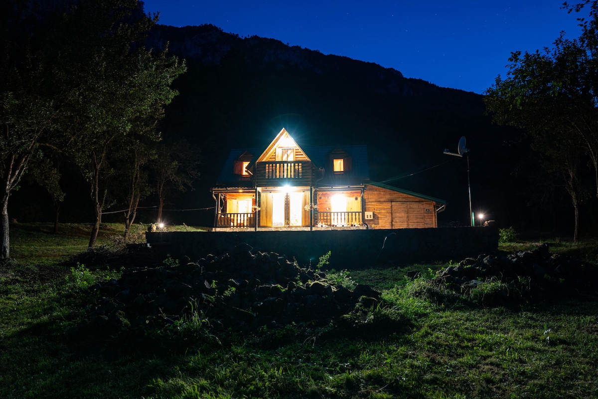 The wooden house is illuminated at night, featuring a welcoming entrance and balcony. Surrounding greenery creates a natural setting, with mountains rising in the background. Soft lights accentuate the structure, inviting a tranquil atmosphere amidst the expansive open space.