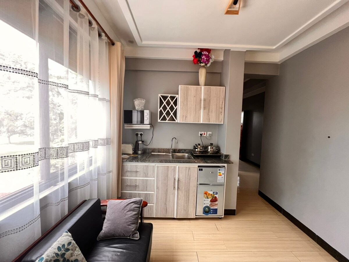 A tidy kitchenette is showcased with a mix of wooden cabinetry and modern appliances. The countertop is equipped with a stove and a microwave, and floral decor adds a touch of color. Ample natural light filters through the sheer curtains, enhancing the space.