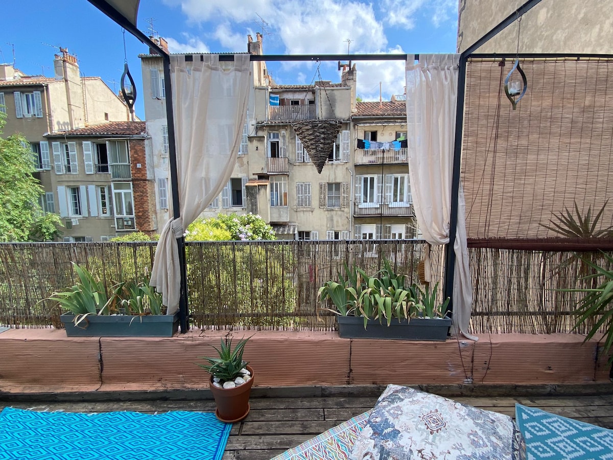 A spacious terrace is shown, featuring a wooden floor and decorated with geometric rugs. Potted plants are arranged along the railing, with sheer curtains framing the view of neighboring buildings and greenery beyond.