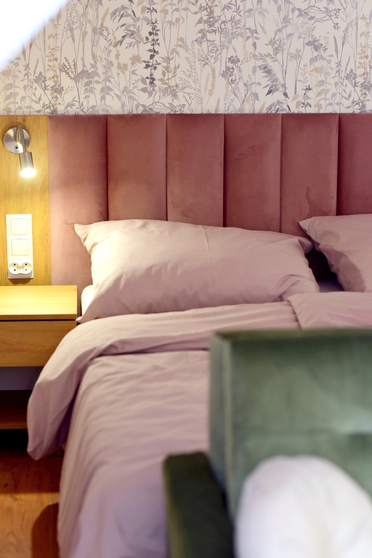 A sleeping area features a soft pink upholstered headboard and cozy bedding. A bedside table is visible with a small light fixture that provides illumination. Decorative floral wallpaper adds a subtle touch to the room's decor.
